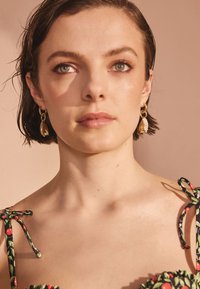 Närbild på en person med kort, mörkt hår som bär guldfärgade örhängen och en blus med blommönster i gröna, röda och svarta färger. Mjukt ljus.