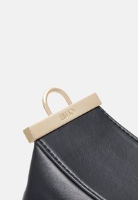 Eckdetails der schwarzen Lederhandtasche mit einem goldfarbenen Verschluss und einer Öse sowie geprägtem Logo. Glatte Textur mit sichtbaren Nähdetails.