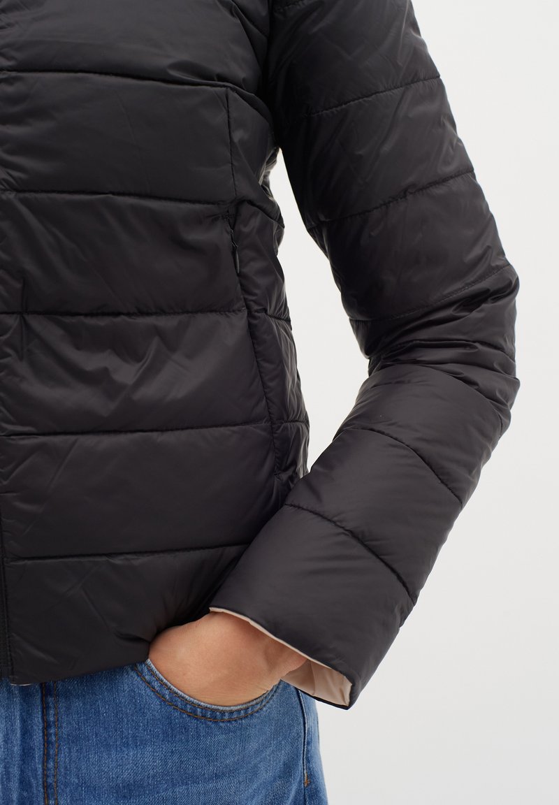 Schwarze gepolsterte Jacke mit Steppdesign, aus glattem Material und leichter Innenausstattung, präsentiert mit einer Hand in der Tasche.