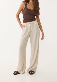 Vrouw in een bruin mouwloos topje, beige broek met wijde pijpen en trekkoord, bruine sandalen en een gouden armband om haar rechterarm.