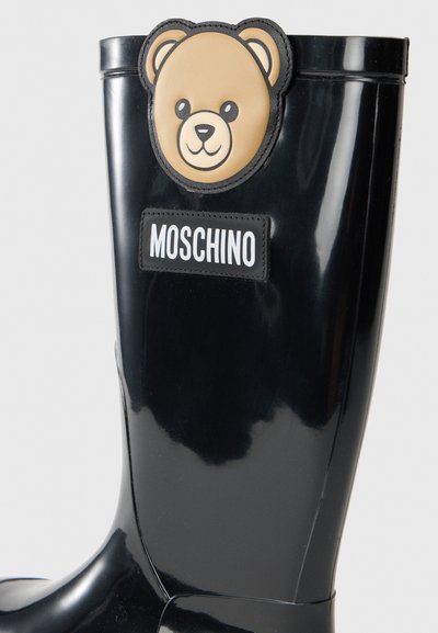 Botte de pluie en caoutchouc noir avec un patch en forme de tête d'ours et le logo MOSCHINO. Texture lisse avec un haut arrondi et une finition brillante.