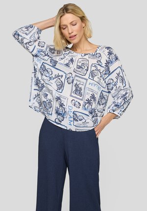 Frau mit kurzen blonden Haaren, die eine weiße und blaue Bluse mit tropischem Muster und dunkle, weite Hosen vor einem einfarbigen grauen Hintergrund trägt.