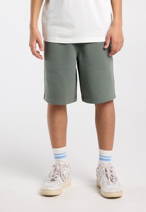 Personne portant un short olive jusqu'aux genoux, des baskets blanches avec des chaussettes blanches à rayures bleues, se tenant contre un fond clair uni.