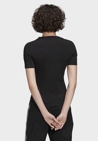 En person med lockigt hår bär en åtsittande svart kortärmad skjorta och svarta byxor med vita ränder, stående med händerna lutade bakom ryggen.