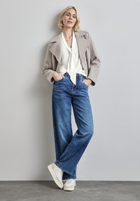 Hellgrauer Wollmischungsmantel, weiße Bluse mit Schleifenkragen und weit geschnittene blaue Jeans mit einem ausgewaschenen Look, kombiniert mit weißen Sneakers.