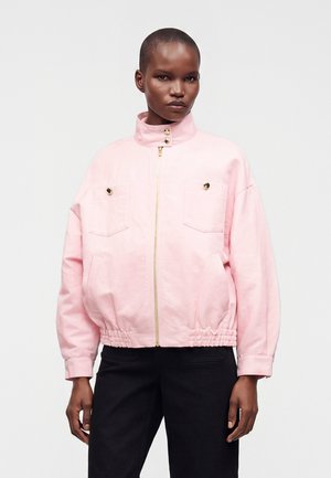 Persona con pelo corto que lleva una chaqueta rosa claro con cremallera, botones dorados y pantalones negros sobre un fondo claro y sencillo.