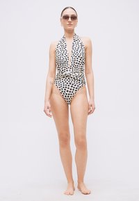 Žena stojaca bosá, oblečená v bielych plavkách s čiernymi bodkami a halter krkom, so slnečnými okuliarmi a náušnicami na bielom pozadí.