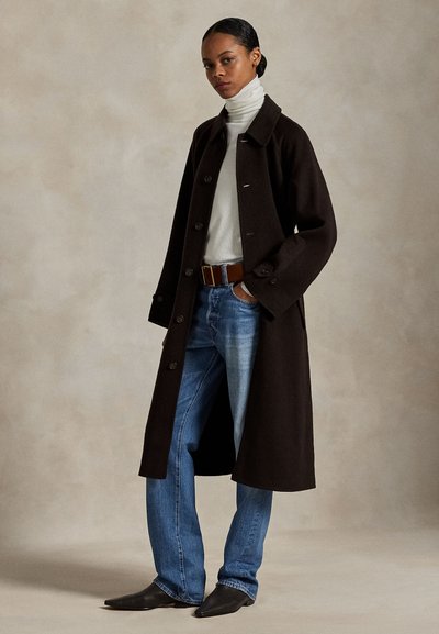 Nő sötét hosszú kabátban, fehér garbóban, kék farmernadrágban barna övvel és fekete hegyes csizmában, semleges háttér előtt áll.