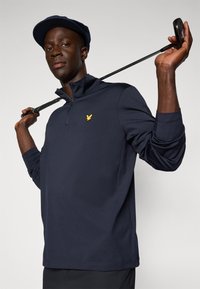 Marineblauer Langarm-Polo mit Reißverschlusskragen und schlanker Passform, mit einem goldenen Adler-Logo auf der Brust. Der Stoff wirkt glatt und leicht.