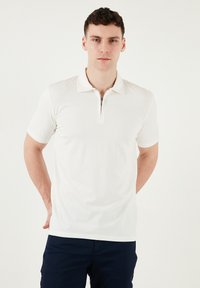 Weißes Polohemd mit kurzen Ärmeln, Reißverschlusskragen, glatter Textur und tailliertem Design, kombiniert mit dunkelblauen Hosen.