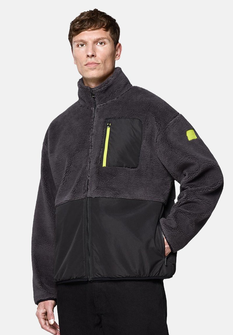 Ciemnoszara polarowa kurtka z czarną dolną częścią, wysokim kołnierzem, zamkiem błyskawicznym z przodu oraz żółtą kieszenią na zamek błyskawiczny na klatce piersiowej.