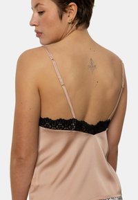 Camisole en satin rose clair avec des bretelles régler. La garniture en dentelle noire au niveau du col présente un motif floral. Le dos affiche un tatouage discret.