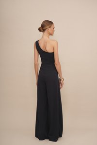 Vrouw met een bun, gekleed in een zwarte één-schouder jumpsuit met brede benen, gouden oorbellen en armbanden, staand tegen een beige achtergrond.