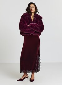 Veste courte en fausse fourrure bourgogne avec des larges manches sur une longue robe en velours bourgogne dotée d'un ourlet en dentelle, associée à des escarpins pointus assortis.