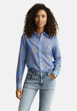 Frau mit lockigem Haar, die ein blaues Hemd mit goldener Kettenmuster und helle Jeans trägt, steht mit einer Hand in der Tasche.