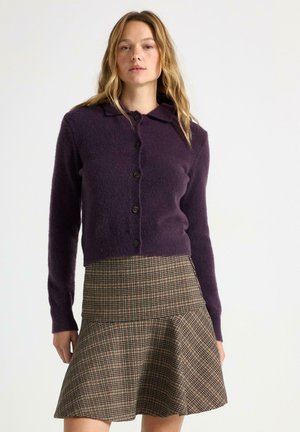 Cardigan in lana viola con collo a bavero e chiusura con bottoni, abbinato a una gonna svasata in tessuto a fantasia scozzese nei toni tenui. Dettagli del tessuto testurizzato visibili.