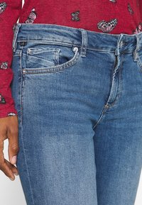 Blue denim jeans with a smooth texture, featuring a mid-rise waist, front pockets, and copper stitching accents.