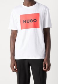 Camiseta de algodón blanca con un gran gráfico rectangular rojo que presenta "HUGO" en letras negras en negrita. Manga corta, cuello redondo.