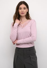 Hosszú barna hajú nő, rózsaszín, hosszú ujjú felsőt visel csipkés dekoltárral és sötét csíkos nadrággal, egy fehér háttér előtt áll.