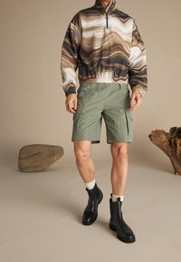 Lynlåsjakke med brunt, hvidt og sort abstrakt mønster; kombineret med olivengrønne shorts med cargolommer; sorte ankelstøvler.