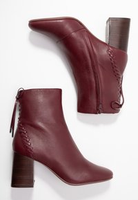 Botas de tobillo de cuero burdeos con puntera afilada, superficie texturizada, cremallera lateral y un tacón bloque de madera. El detalle de cordones laterales añade interés.