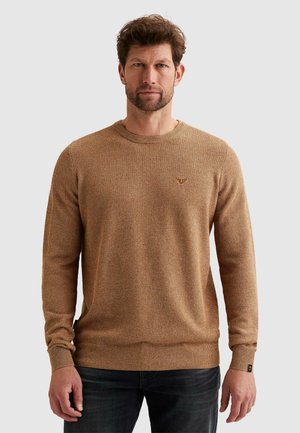 Muž oblečený v textúrovanom svetlohnedom svetri s okrúhlym výstrihom a tmavých džínsach, stojaci pred jednoduchým svetlosivým pozadím.