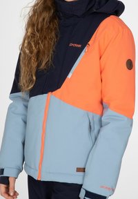Farbblockierte Winterjacke mit navyblauen, orangefarbenen und hellblauen Paneelen, aus synthetischem Material, mit Reißverschluss und Logodetail am Ärmel.