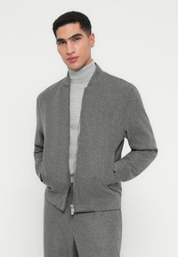 Uomo in maglione a collo alto grigio chiaro e giacca con zip grigio antracite, pantaloni, davanti a uno sfondo bianco semplice.