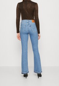 Jean évasé bleu clair à taille haute, avec des surpiqûres contrastantes et une pièce en cuir à l'arrière. Associé à un haut marron ajusté et texturé.