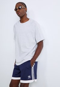 Muž v svetlosivom tričku Adidas, námorníckych modrých šortkách Adidas s bielymi pruhmi a béžových slnečných okuliaroch, stojí proti bielej stene s rukami v vreckách.