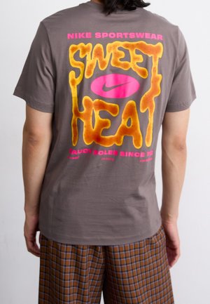 Osoba nosząca szarą koszulkę Nike z dużym żółtym napisem „Sweet Heat” i różowym logo Nike z tyłu, w połączeniu z brązowymi spodnimi w kratę.