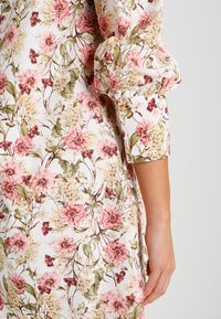 Tissu à motif floral avec des fleurs roses et vertes sur un fond blanc. Détail de manches froncées et un bouton au poignet.