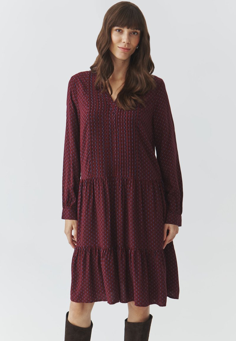 Robe longue bordeaux à manches longues avec décolleté en V, présentant de subtils motifs géométriques, une jupe à volants et des poignets à bouton, fabriquée en tissu léger.