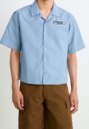 Homme portant une chemise à manches courtes bleu clair avec un logo Puma sur la poitrine et un short cargo marron, debout devant un fond uni.