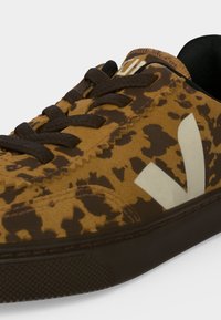 Gros plan d'une basket marron et noire tachetée avec des lacets noirs et le logo blanc "V" sur le côté sur un fond gris.
