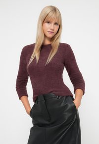 Νεαρή γυναίκα με μακριά ξανθά μαλλιά, φορώντας ένα αφράτο σκούρο μωβ πουλόβερ και μια μαύρη δερμάτινη φούστα, με τα χέρια στις τσέπες, σε ουδέτερο φόντο.