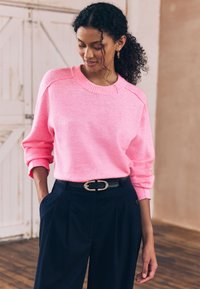 Suéter rosa de punto con cuello acanalado y costuras caídas en los hombros, combinado con pantalones azul marino de talle alto y un cinturón negro con hebilla dorada.