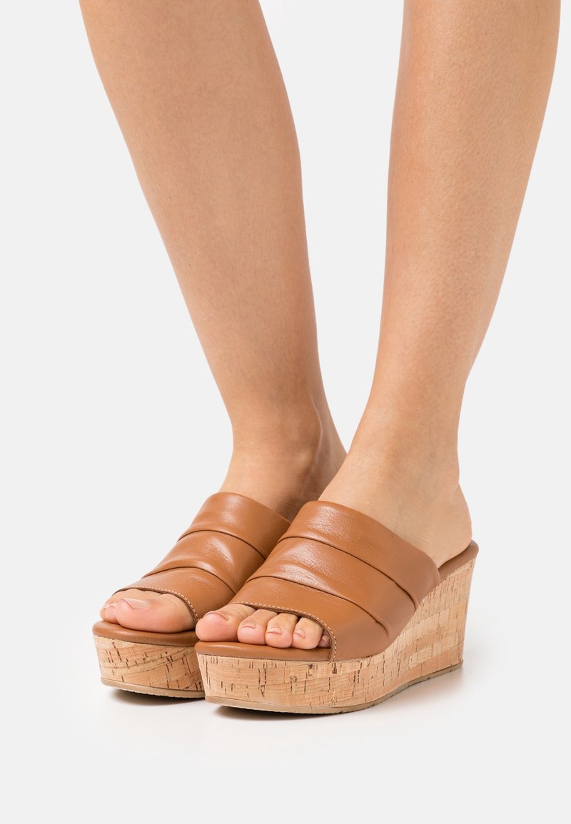 Pieds portant des sandales compensées en cuir marron avec semelles en liège sur fond blanc, montrant les orteils et le bas des jambes.