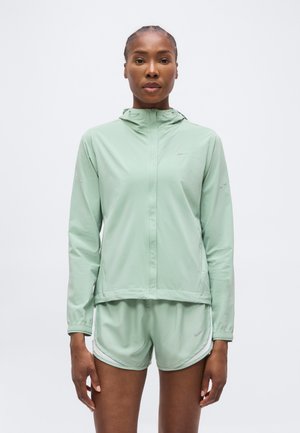 Frau trägt hellgrüne Kapuzenjacke und passende Shorts, steht vor weißem Hintergrund, blickt nach vorne.