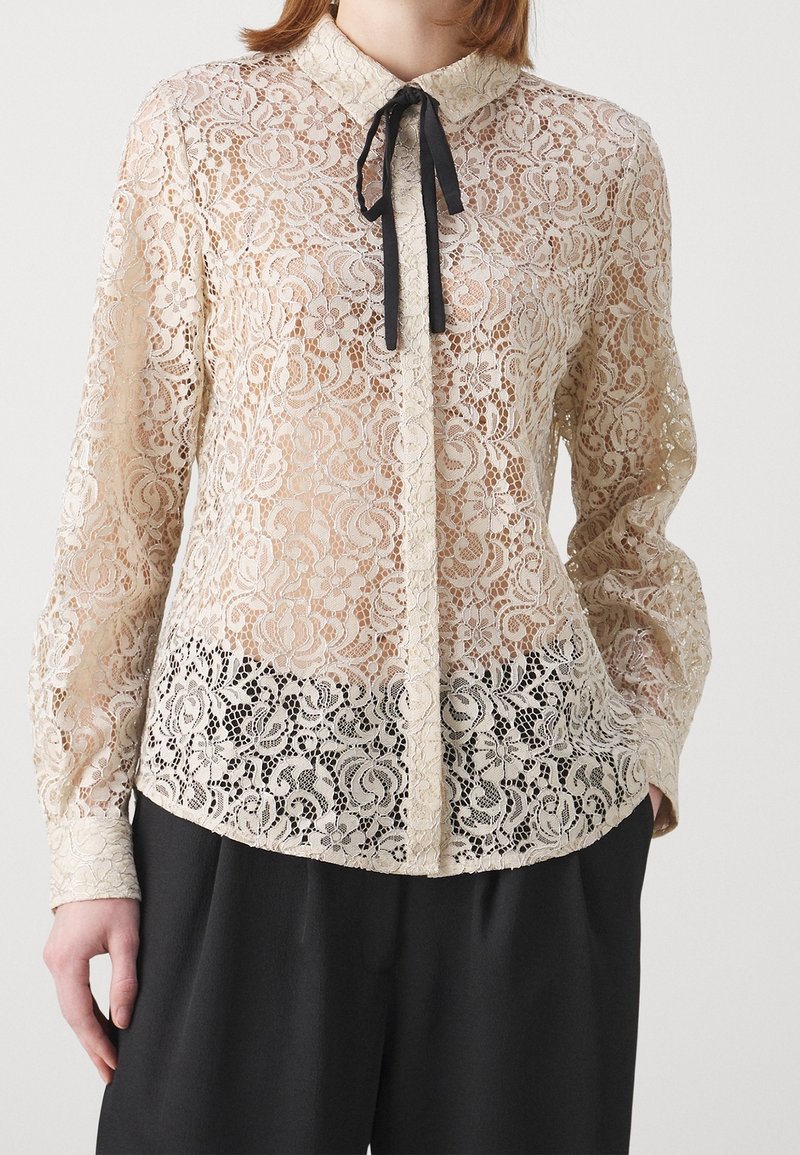 Blouse longue en dentelle crème avec motif floral et ruban noir noué au col, portée avec un pantalon noir.