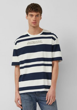 Giovane uomo con maglietta a righe blu navy e bianche con la scritta "Feel Observe Grow Rethink Reflect & Act" e jeans blu, in piedi davanti a uno sfondo grigio.