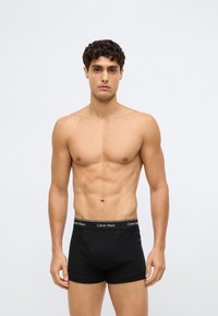 Homem sem camisa com cabelo encaracolado, vestindo boxers pretos da Calvin Klein, em pé contra um fundo cinza claro liso.