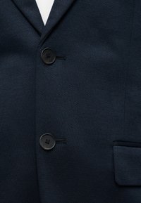 Tmavomodrý sako so štruktúrovanou látkou, zapnuté na dva čierne gombíky, s vykrojeným klopovým golierom a predným náprsníkom s klapkou na pravej strane.