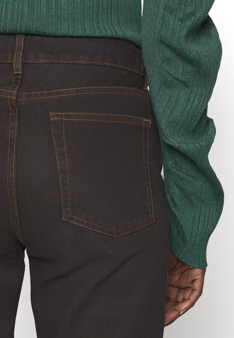 Braune Jeans aus Denim mit kontrastierenden gelben Nähten, versehen mit einer Gesäßtasche und getragen mit einem strukturierten, grünen Rippenpullover.