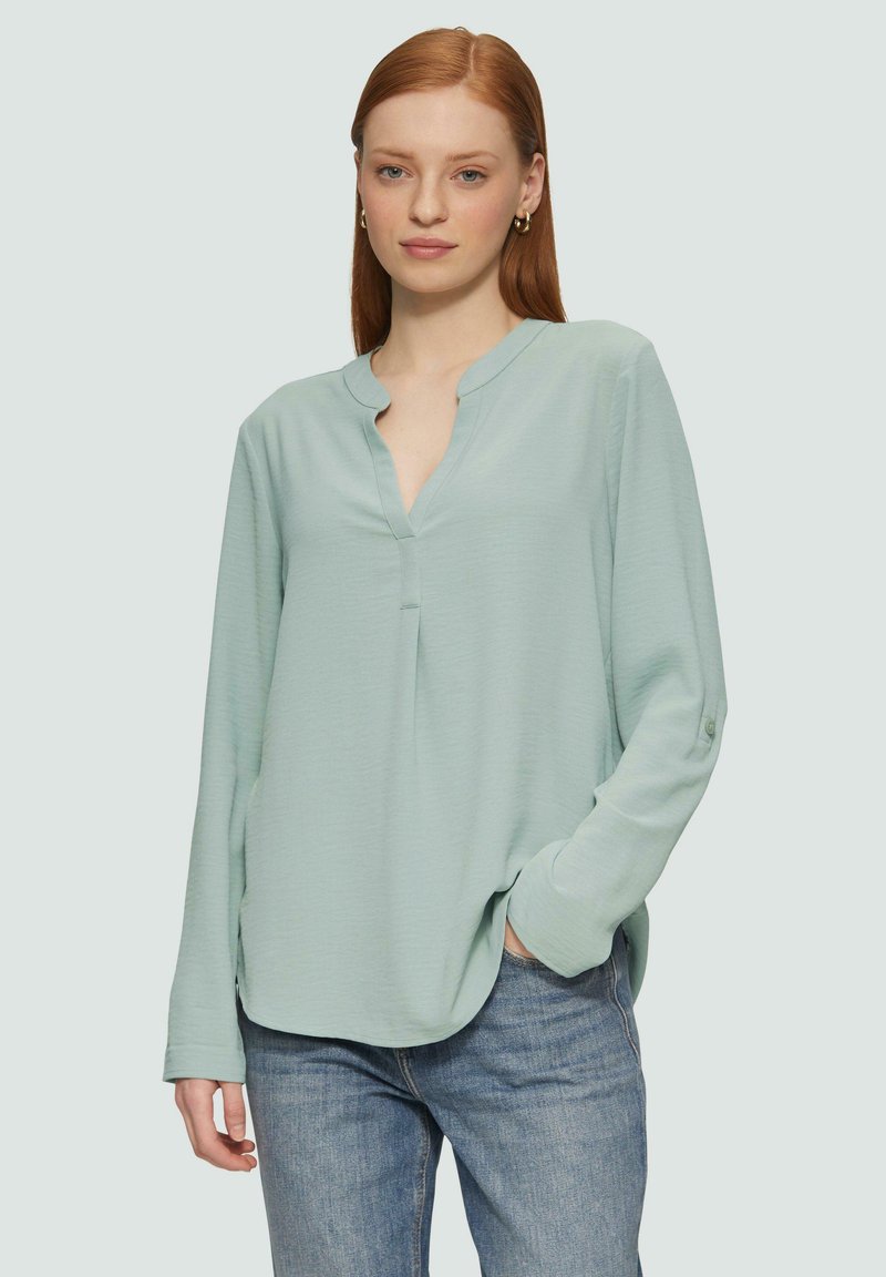 Blouse bleu clair à manches longues avec un col à revers, offrant une coupe décontractée et des poches latérales, fabriquée à partir d'un tissu doux et texturé.