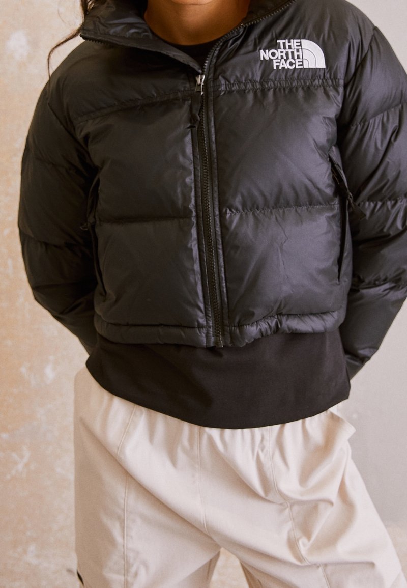 Svart puffjacka med frontdragkedja, hög krage och The North Face-logotyp. Har kviltade sektioner och en avslappnad passform.