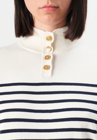 Maglione bianco a maglia con strisce orizzontali blu navy, colletto alto e quattro bottoni dorati sul pattino. Texture liscia.
