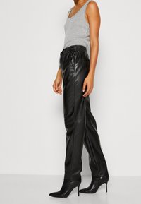 Zwarte leren broek met een relaxed fit en elastische tailleband, gecombineerd met een grijze tanktop en puntige zwarte hakken, die een slanke silhouet benadrukken.