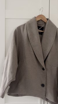 Beige, übergroße Jacke aus weichem Stoff mit Schalkragen, Knöpfen vorne und zwei Seitentaschen. Glatte Textur mit auffälligen Nähten.