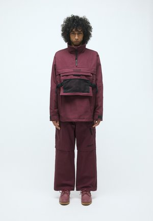 Burgunderfarbige Outdoor-Jacke mit Viertel-Reißverschluss und Netz-Fronttasche, kombiniert mit weiten burgunderfarbenen Hosen und burgunderfarbenen Schuhen mit strukturierten Sohlen.